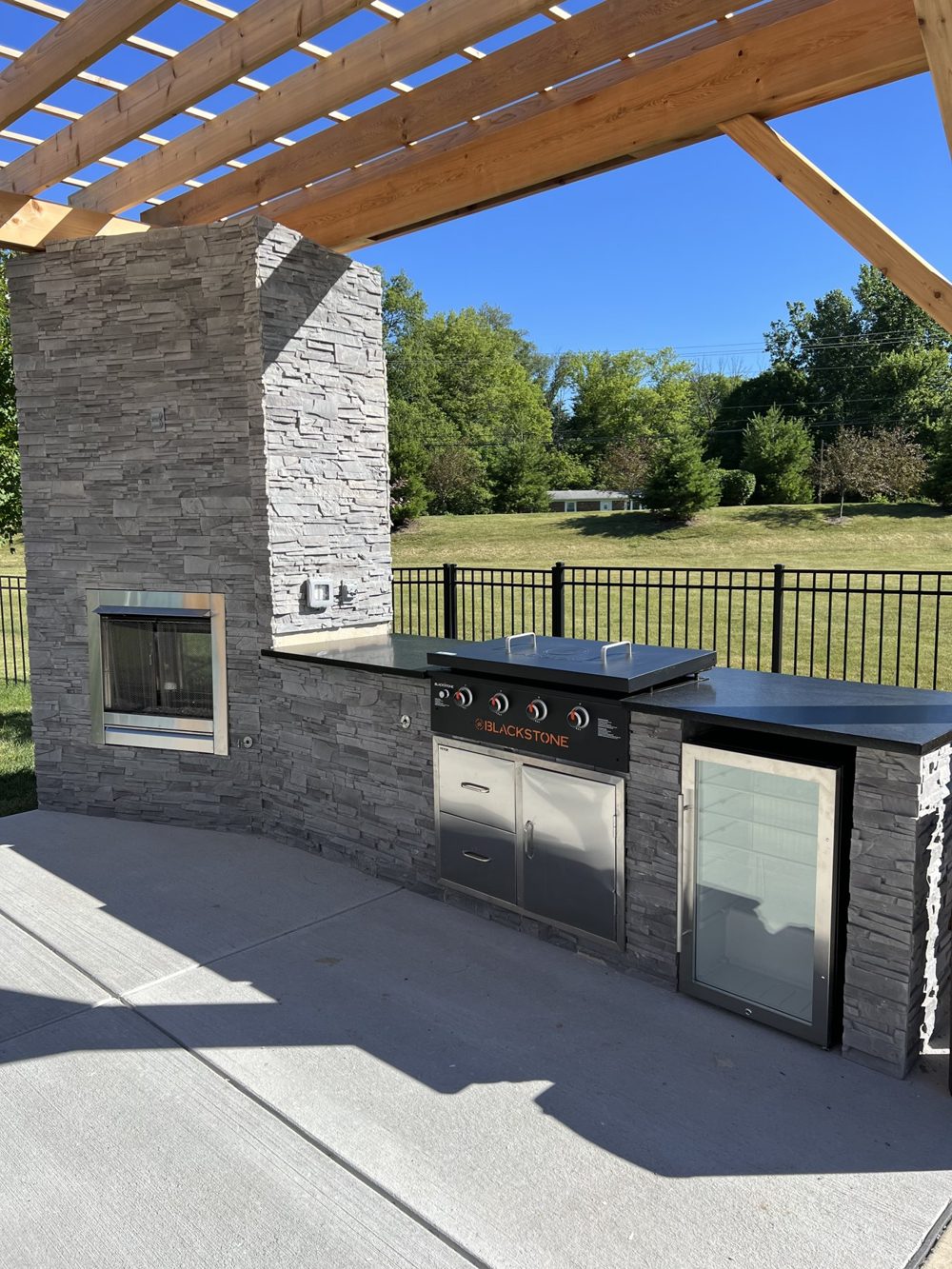 Stone outdoor kitchen under a timber pergola beside a fenced yard